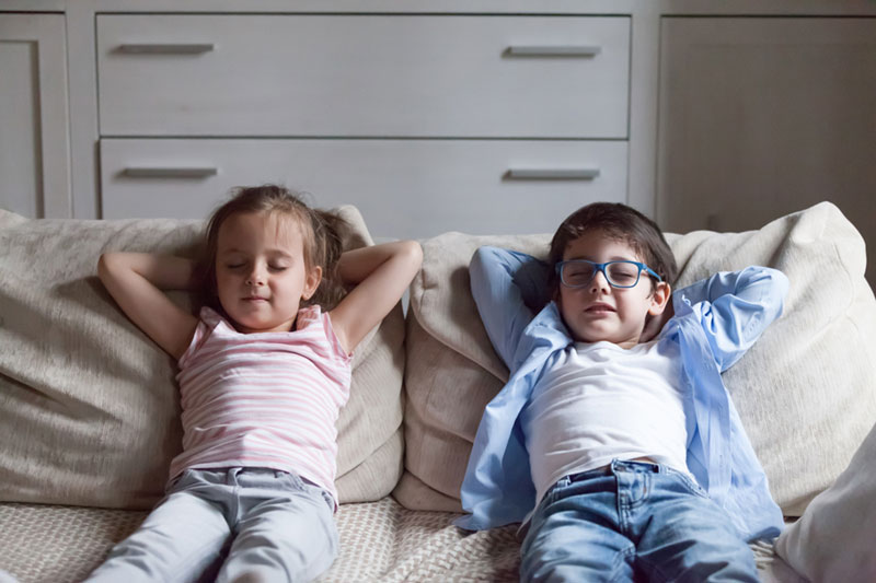 Silent preschool sister and brother sitting on sofa in living room at home. Siblings closing eyes have break breath fresh air putting hands behind heads relaxing thinking.